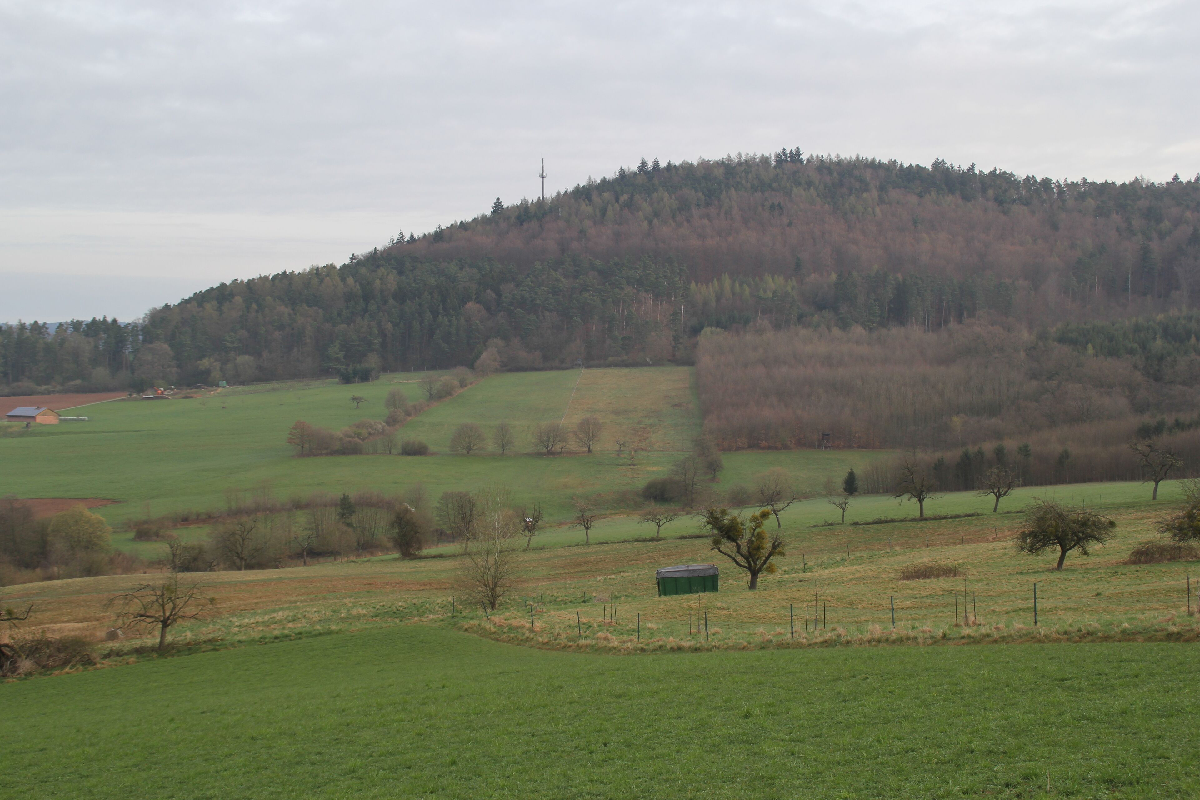 A rural landscape with rolling hills, a forested hilltop, and a small green building in the foreground.