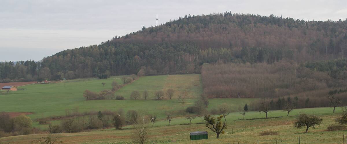 A rural landscape with rolling hills, a forested hilltop, and a small green building in the foreground.