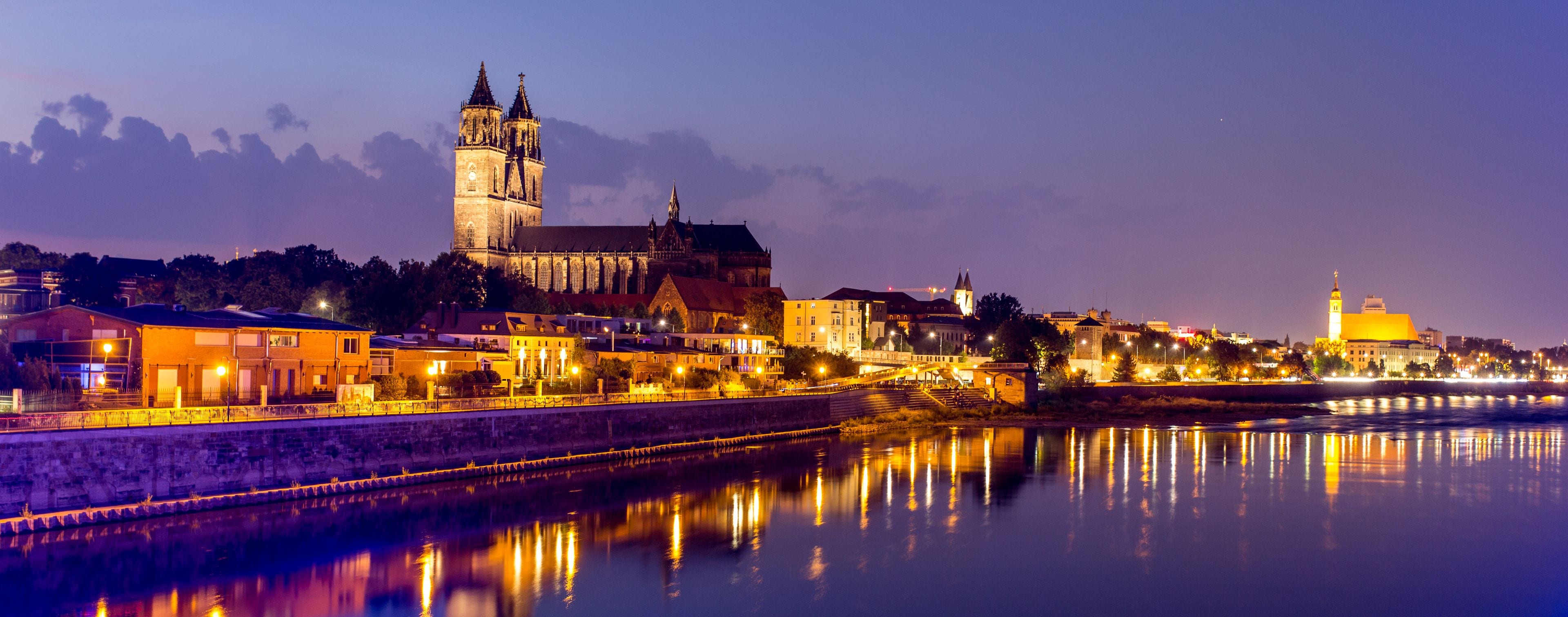 A historic cityscape with a prominent cathedral and tower, a river reflecting the lights, and a bridge connecting the two sides.