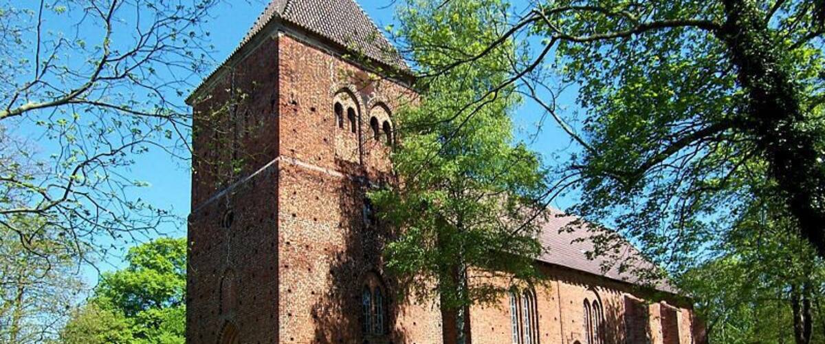 A brick church with a tall tower and arched windows, surrounded by lush green trees.