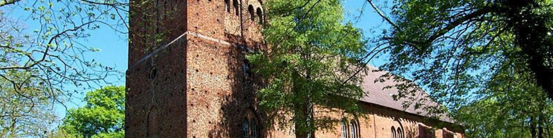 A brick church with a tall tower and arched windows, surrounded by lush green trees.