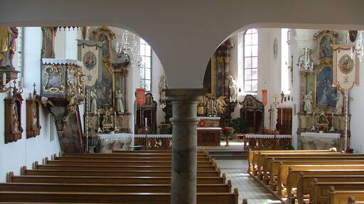 Interior de una iglesia ornamentada con bancales de madera, estatuas religiosas y vidrieras.