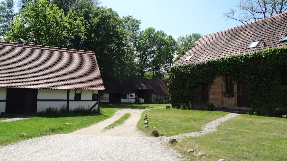 Un cadre rural avec des bâtiments traditionnels, un chemin de gravier et une végétation luxuriante.