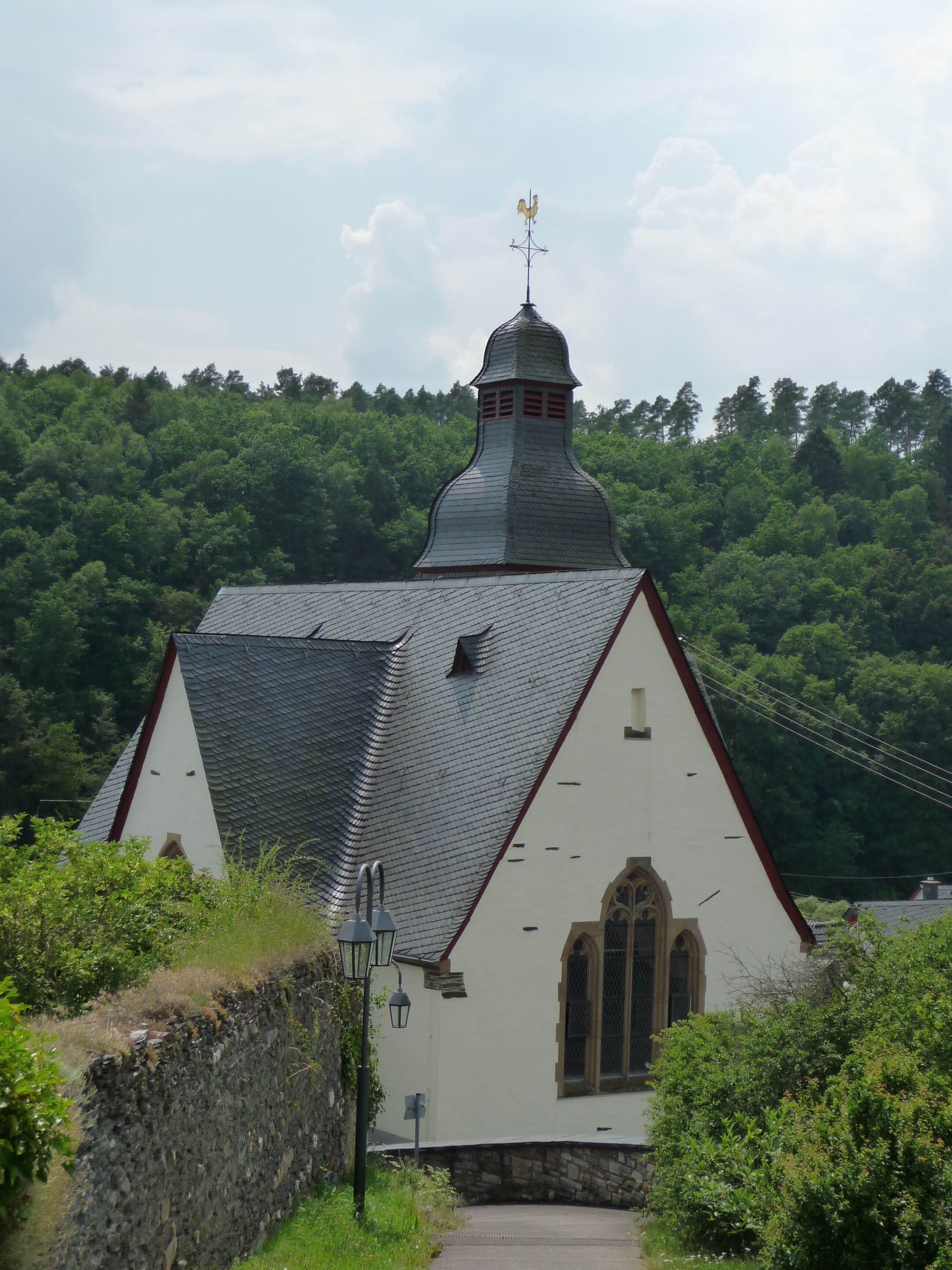 A church with a pointed roof and a weather vane on top, surrounded by lush greenery.