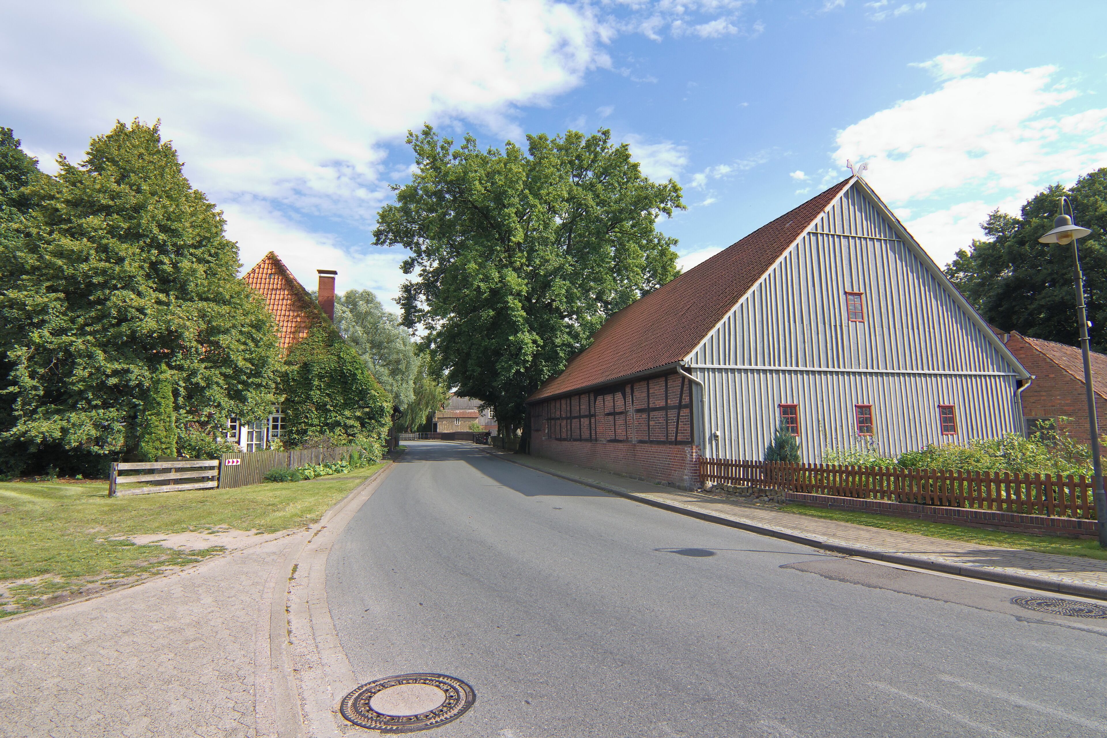 A quiet street with a large white barn, a red-roofed house, and lush green trees.