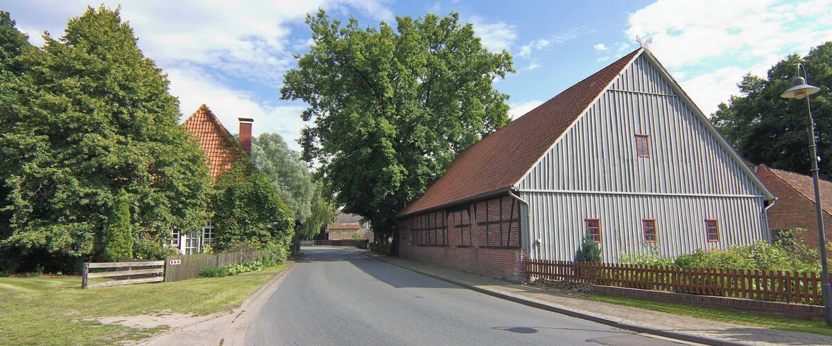 A quiet street with a large white barn, a red-roofed house, and lush green trees.