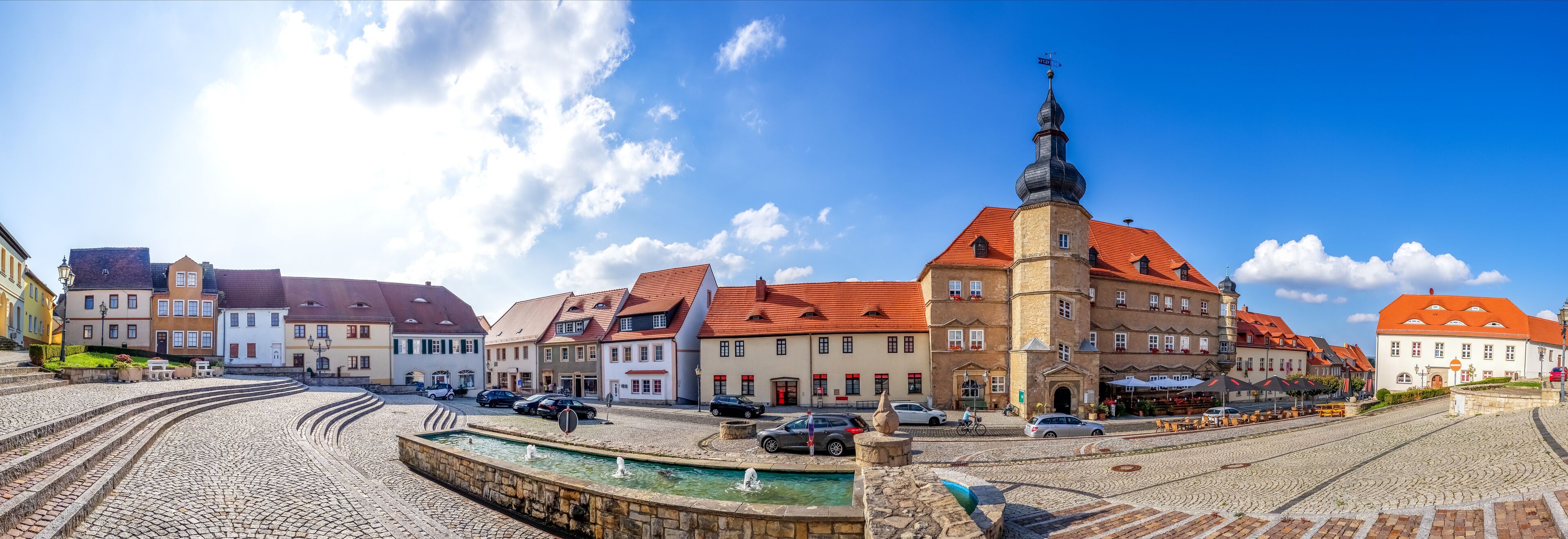 A cobblestone square with historic buildings, a church tower, and a fountain.