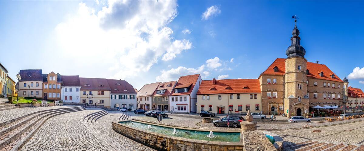 A cobblestone square with historic buildings, a church tower, and a fountain.