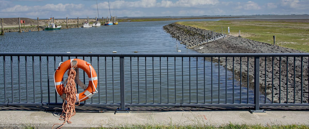 A calm body of water with a lifebuoy hanging on a railing, boats docked on the shore, and a clear sky.