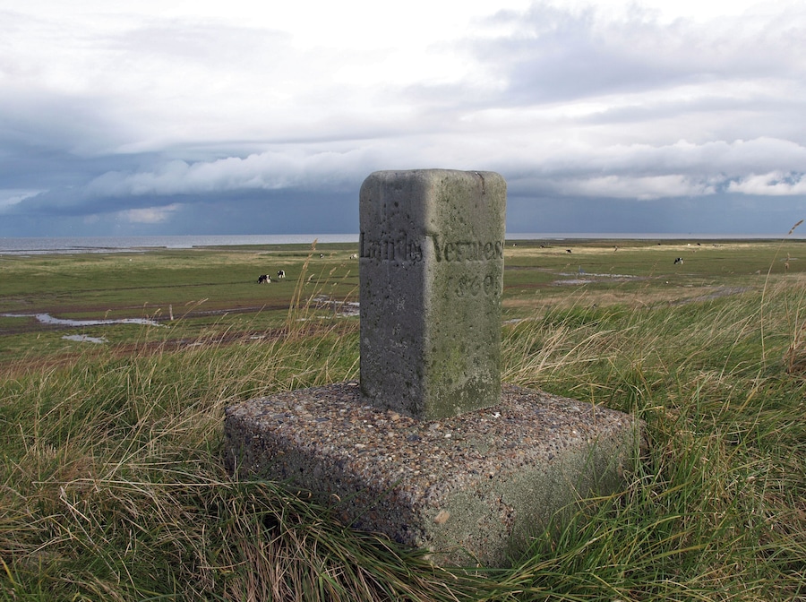 Monumento de piedra con inscripciones en un campo de césped, con vacas al fondo.