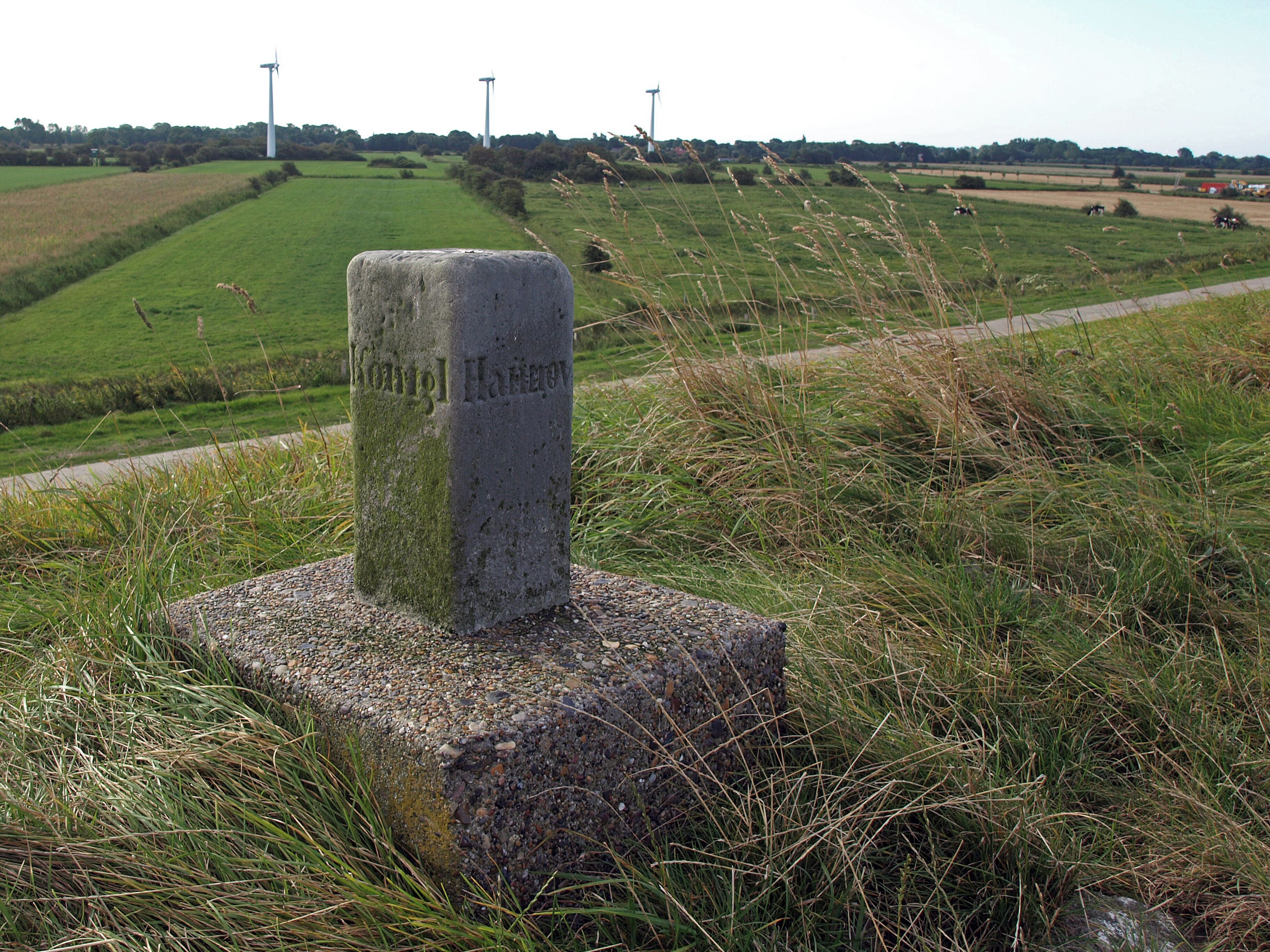 A stone monument with inscriptions in a rural landscape with fields and wind turbines in the background.