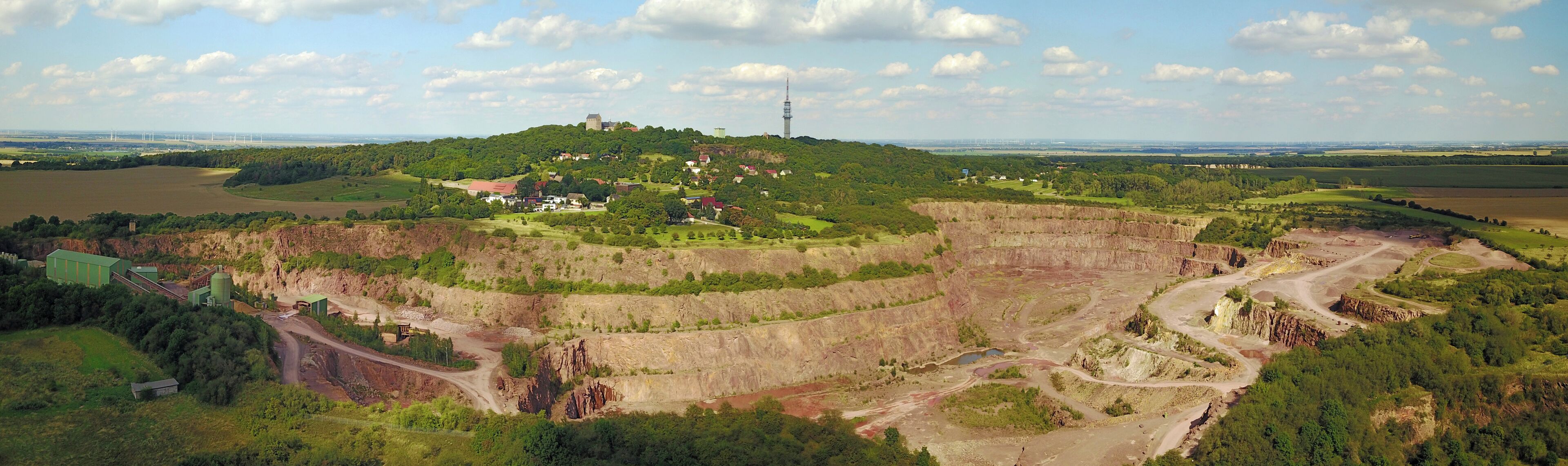 A mining site with terraced excavation areas, a hilltop building, and a wind farm in the distance.