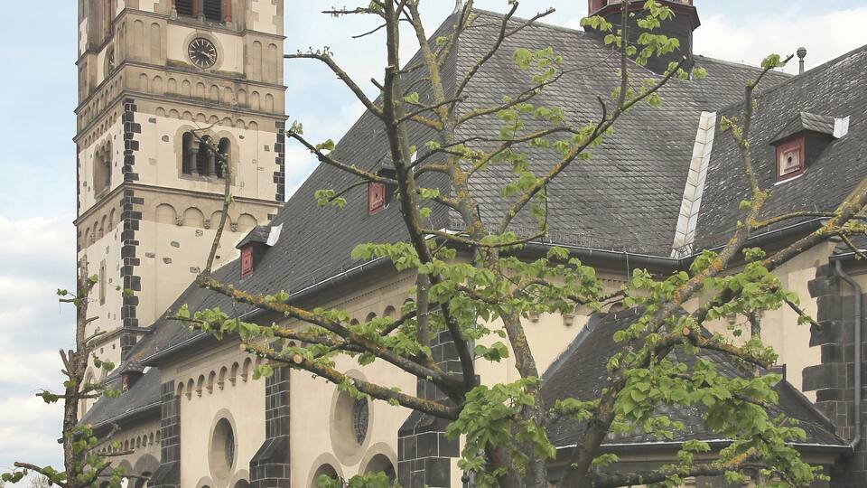 A historic church with a tall bell tower and a smaller spire, surrounded by trees and a parked car.