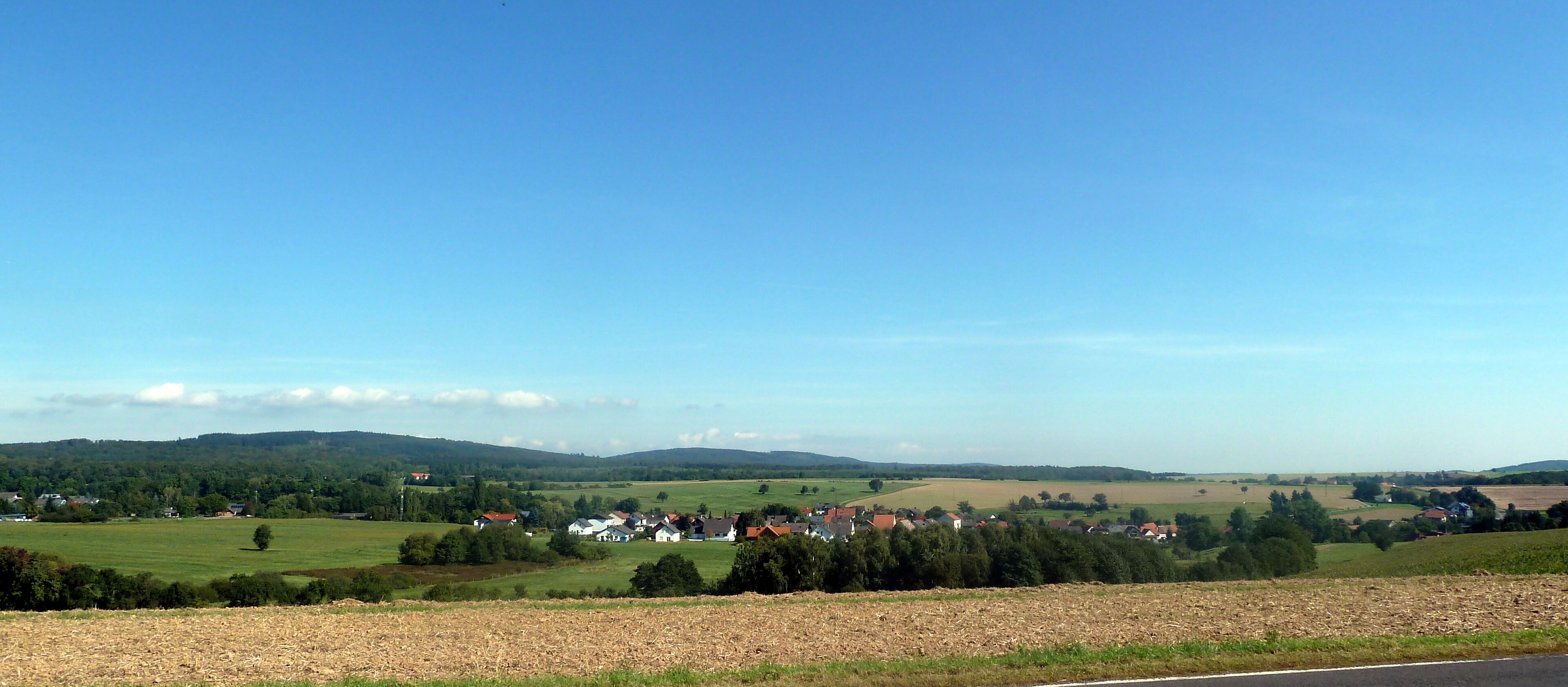 A rural landscape with a clear blue sky, rolling hills, and scattered houses.