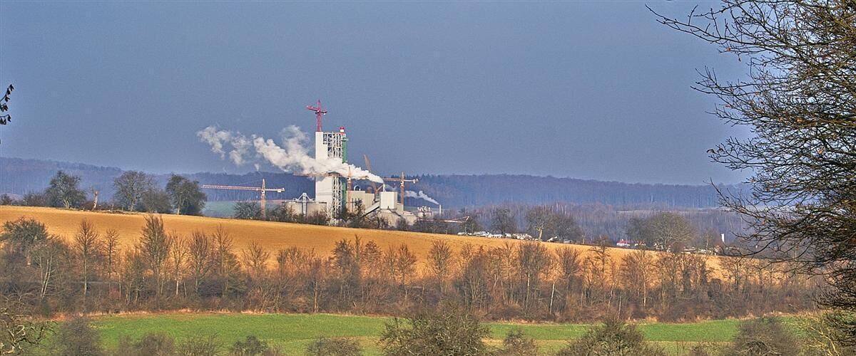 A rural landscape with a large industrial facility emitting smoke, surrounded by fields and trees.