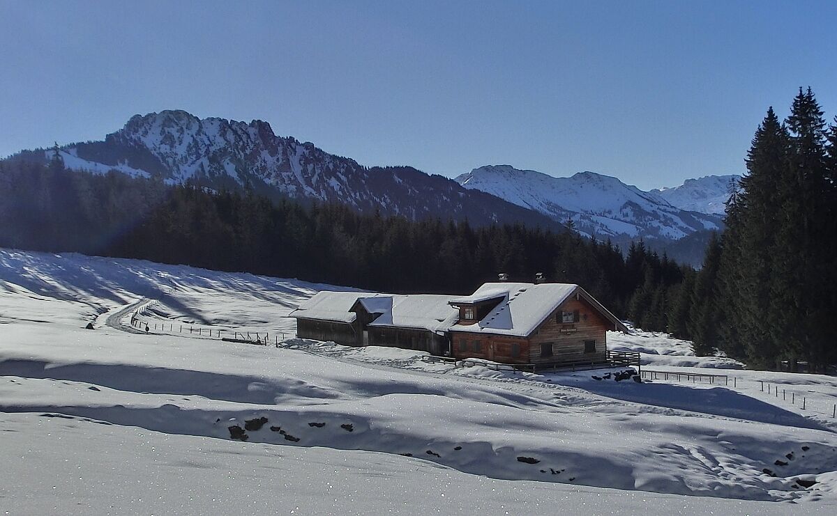 A wooden cabin with a snow-covered roof, surrounded by a snowy landscape and a forested mountain range.