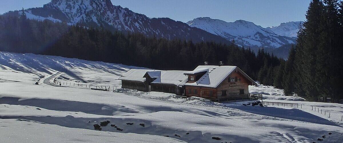 A wooden cabin with a snow-covered roof, surrounded by a snowy landscape and a forested mountain range.