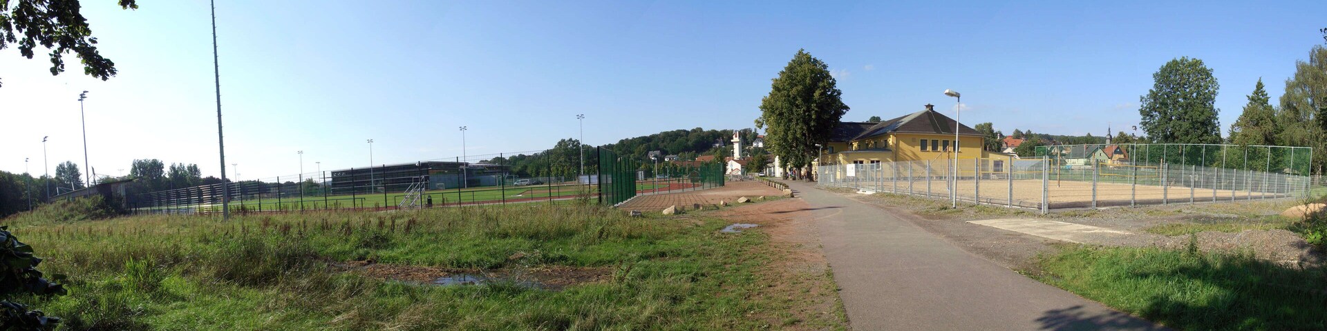 A sports field with a pathway leading to a building, surrounded by fences and trees.