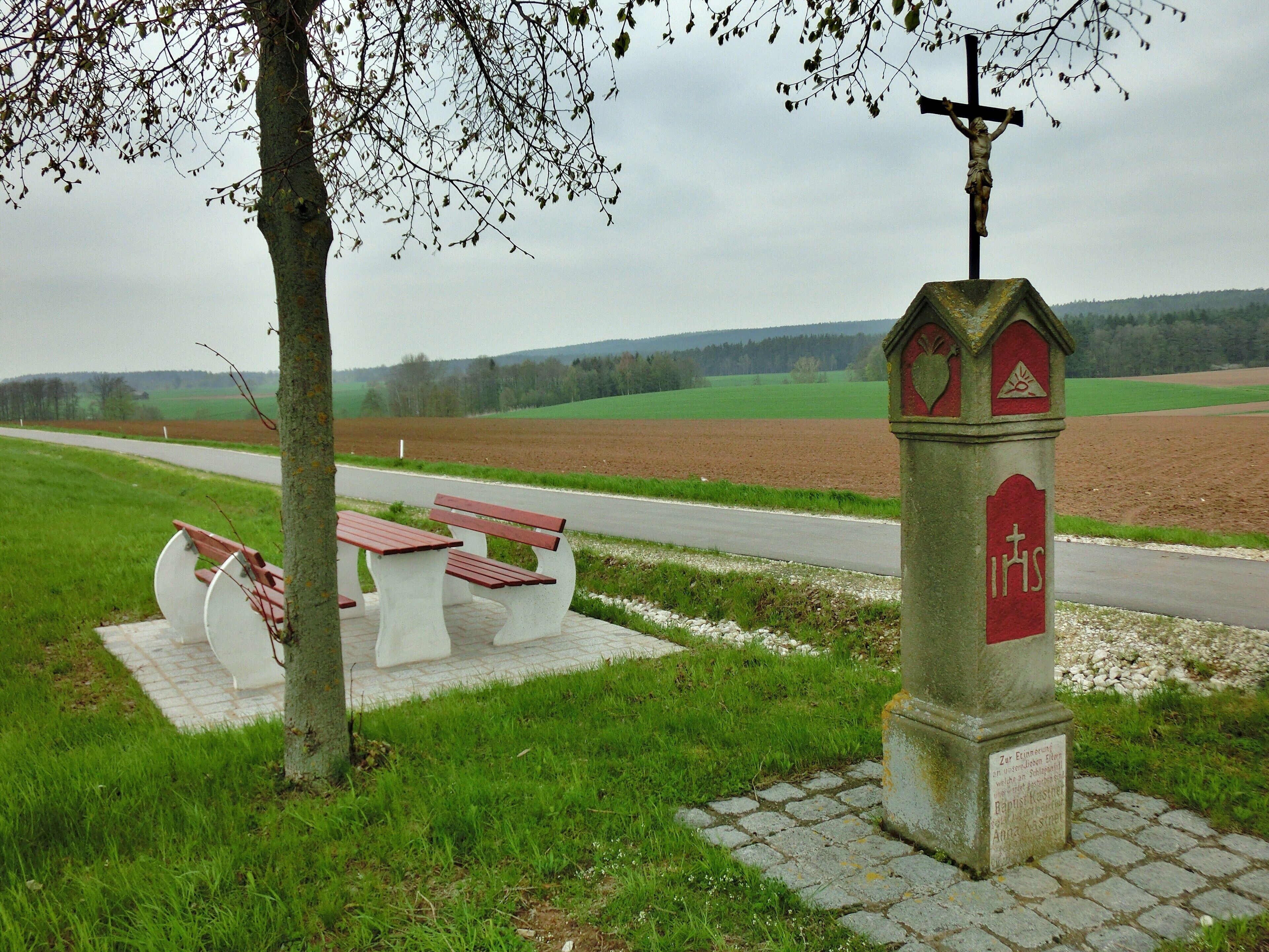 A rural landscape with a cross monument, picnic tables, and a road.