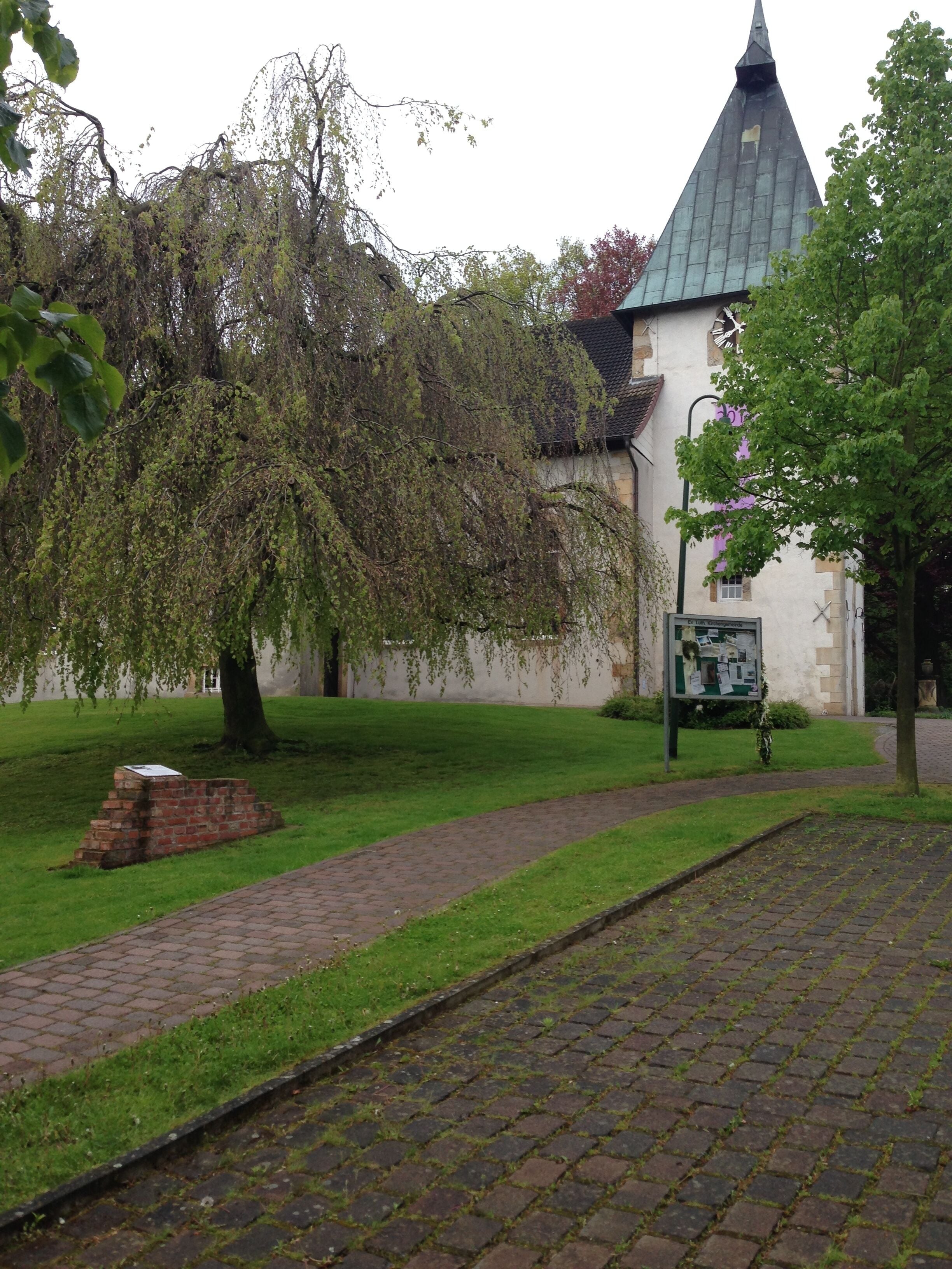 A church with a pointed steeple, a brick wall, and a signboard.