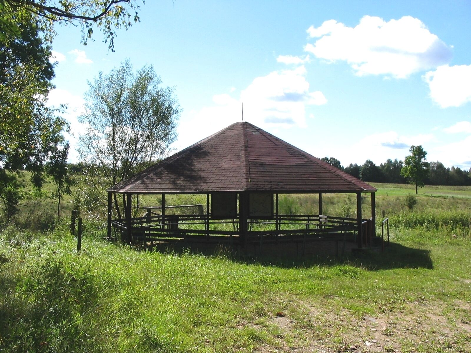A wooden gazebo in a grassy field with trees in the background.