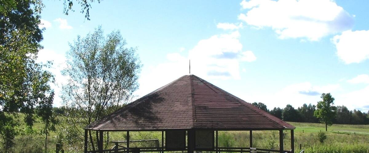 A wooden gazebo in a grassy field with trees in the background.