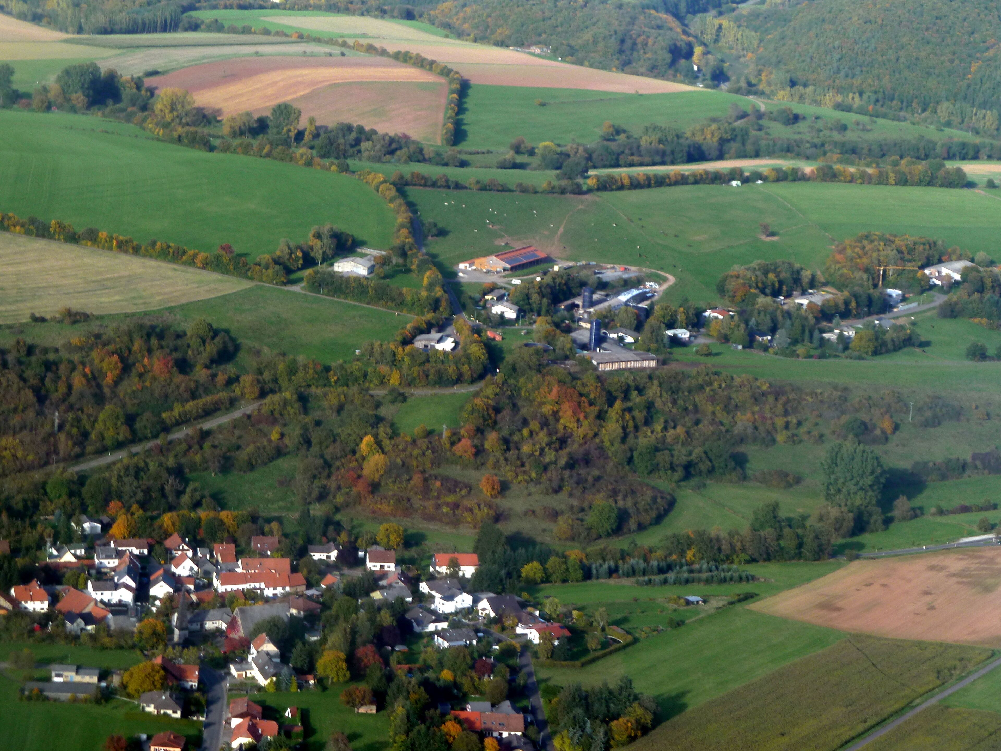 A rural landscape with rolling hills, fields, and a small village with houses and a large industrial building.