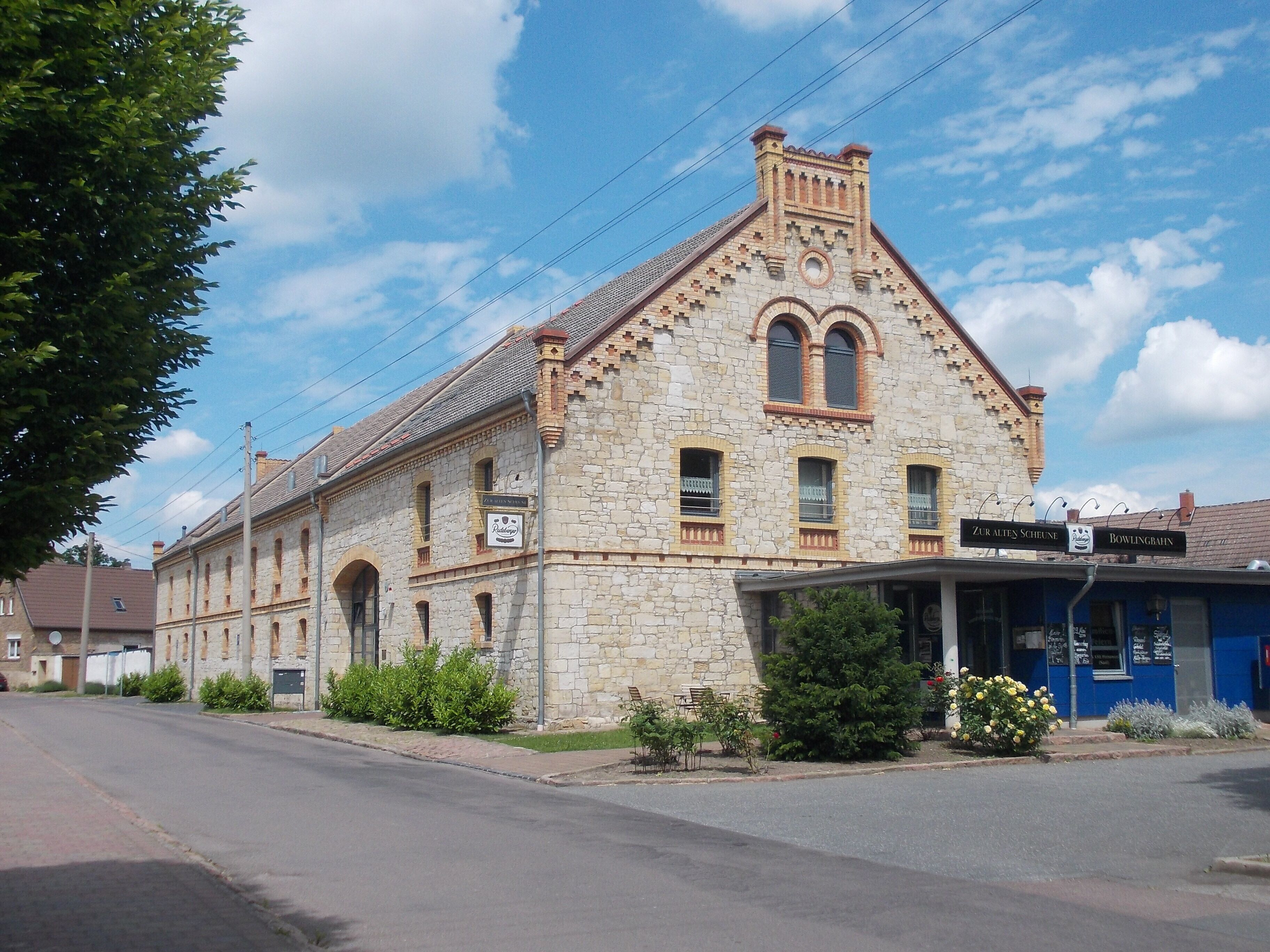 A stone building with arched windows and a sign that reads "Zur Alten Scheune Bowlingbahn."