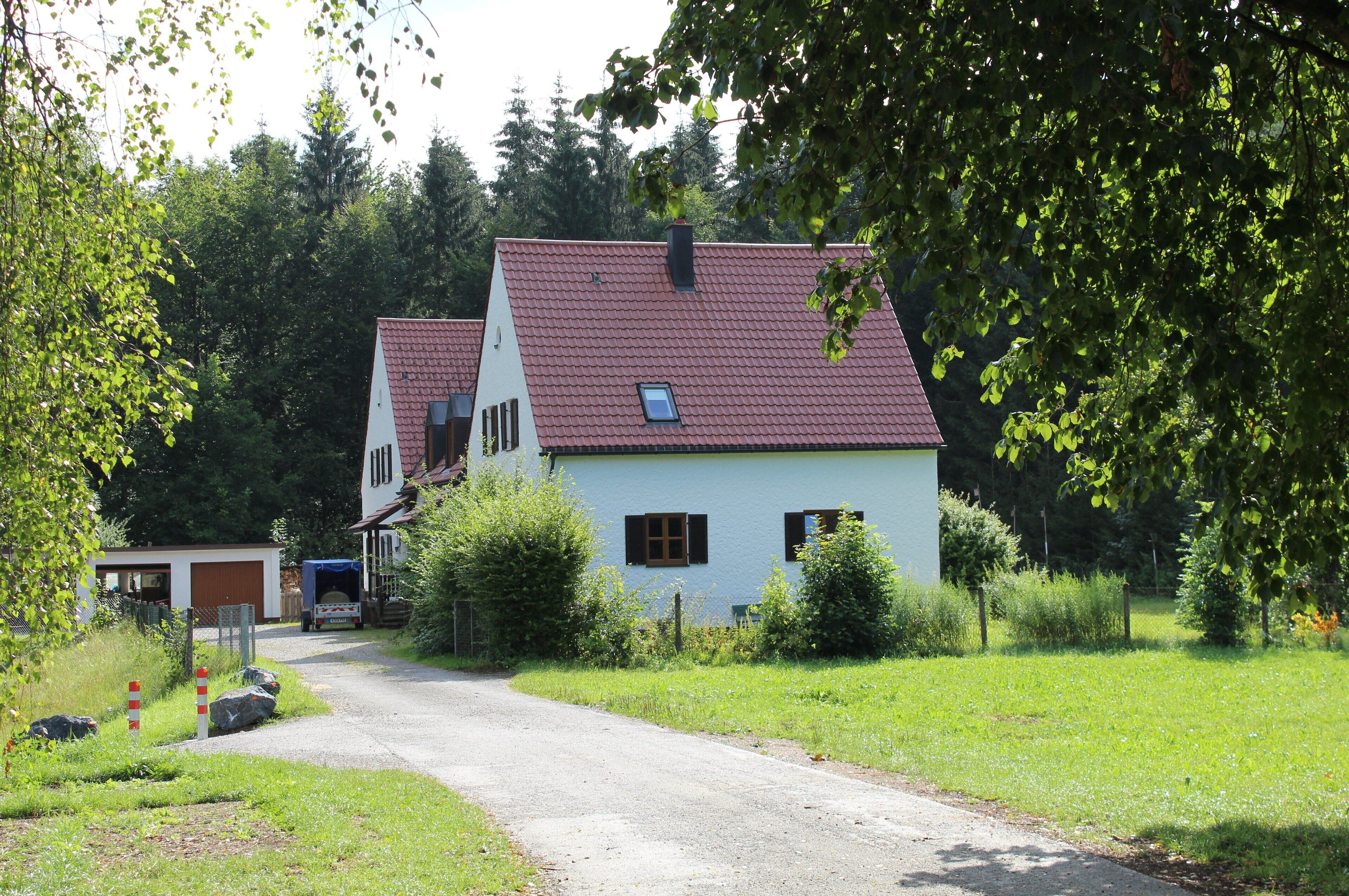 A rural road leading to a white house with a red roof, surrounded by greenery and trees.