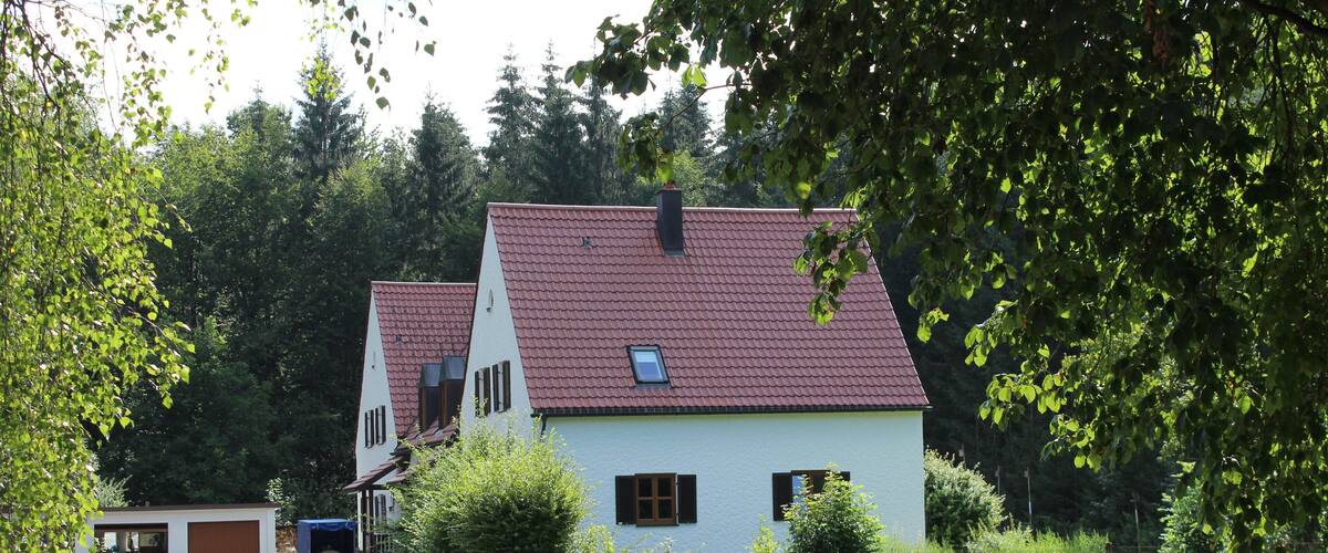 A rural road leading to a white house with a red roof, surrounded by greenery and trees.