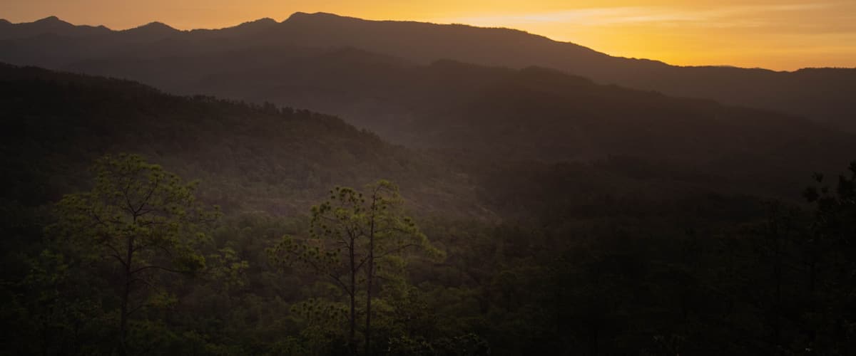 A mountainous landscape at sunset with silhouettes of trees and hills.