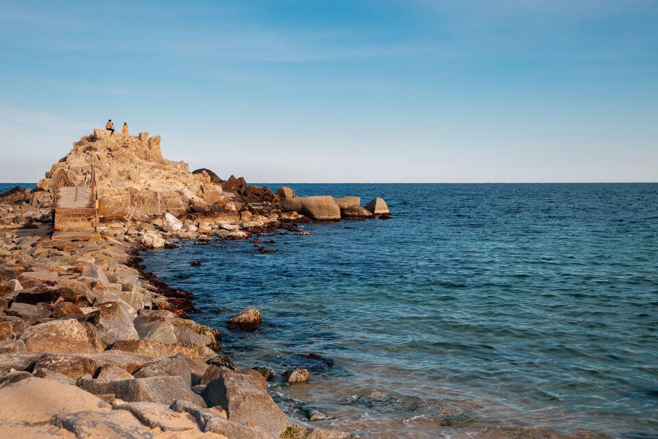 A rocky coastline with a small staircase leading up to a rocky outcrop where two people are standing.