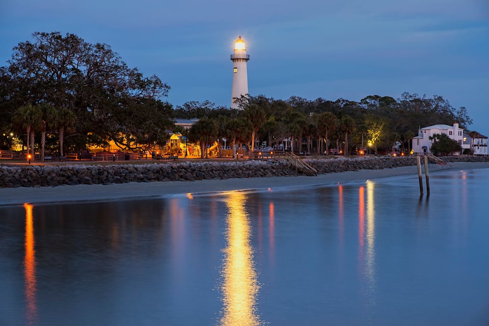 A calm waterfront with a lighthouse, trees, and a stone wall.