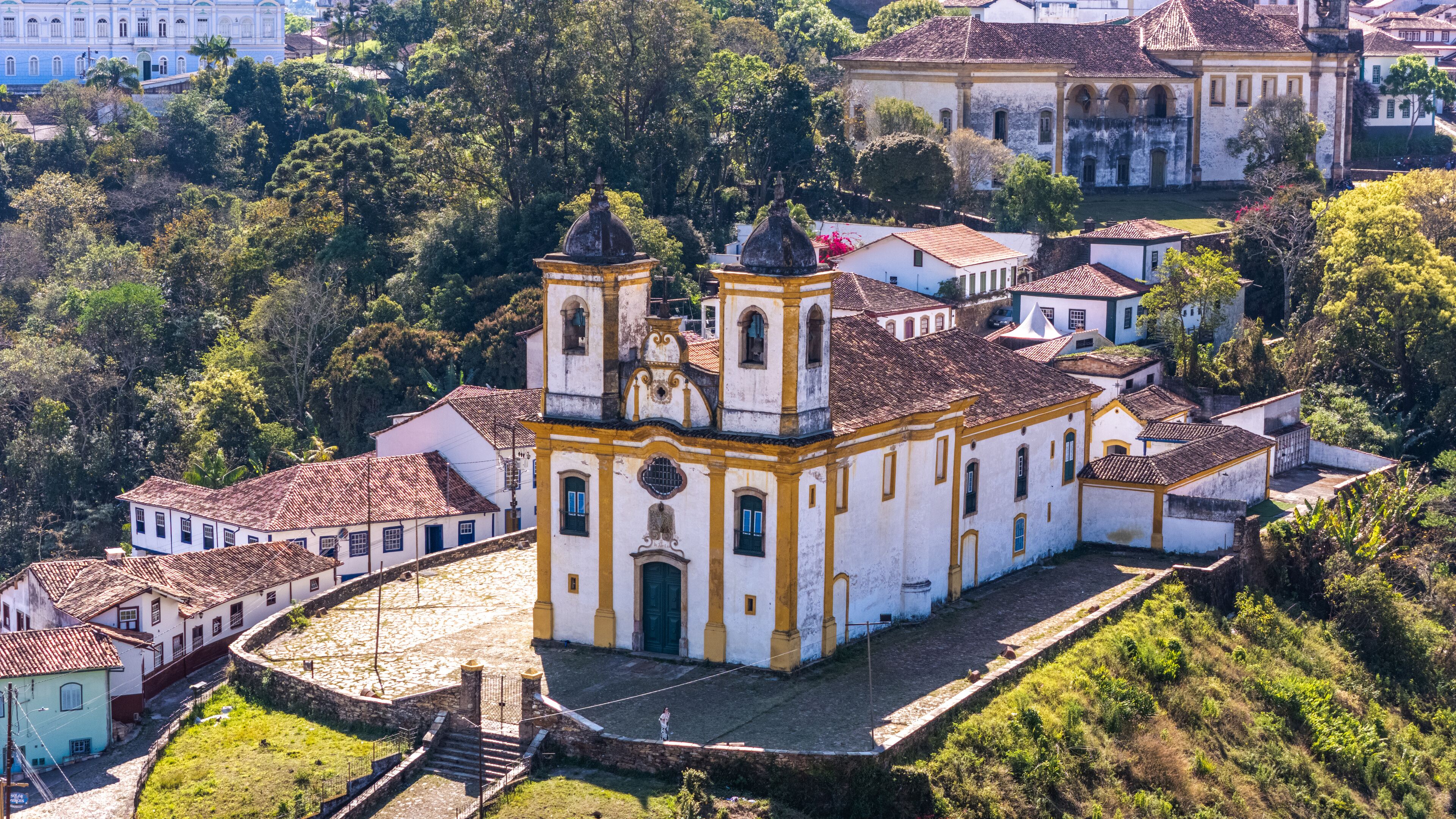 A white church with yellow trim and two bell towers, surrounded by lush greenery and traditional buildings.