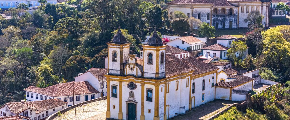 A white church with yellow trim and two bell towers, surrounded by lush greenery and traditional buildings.