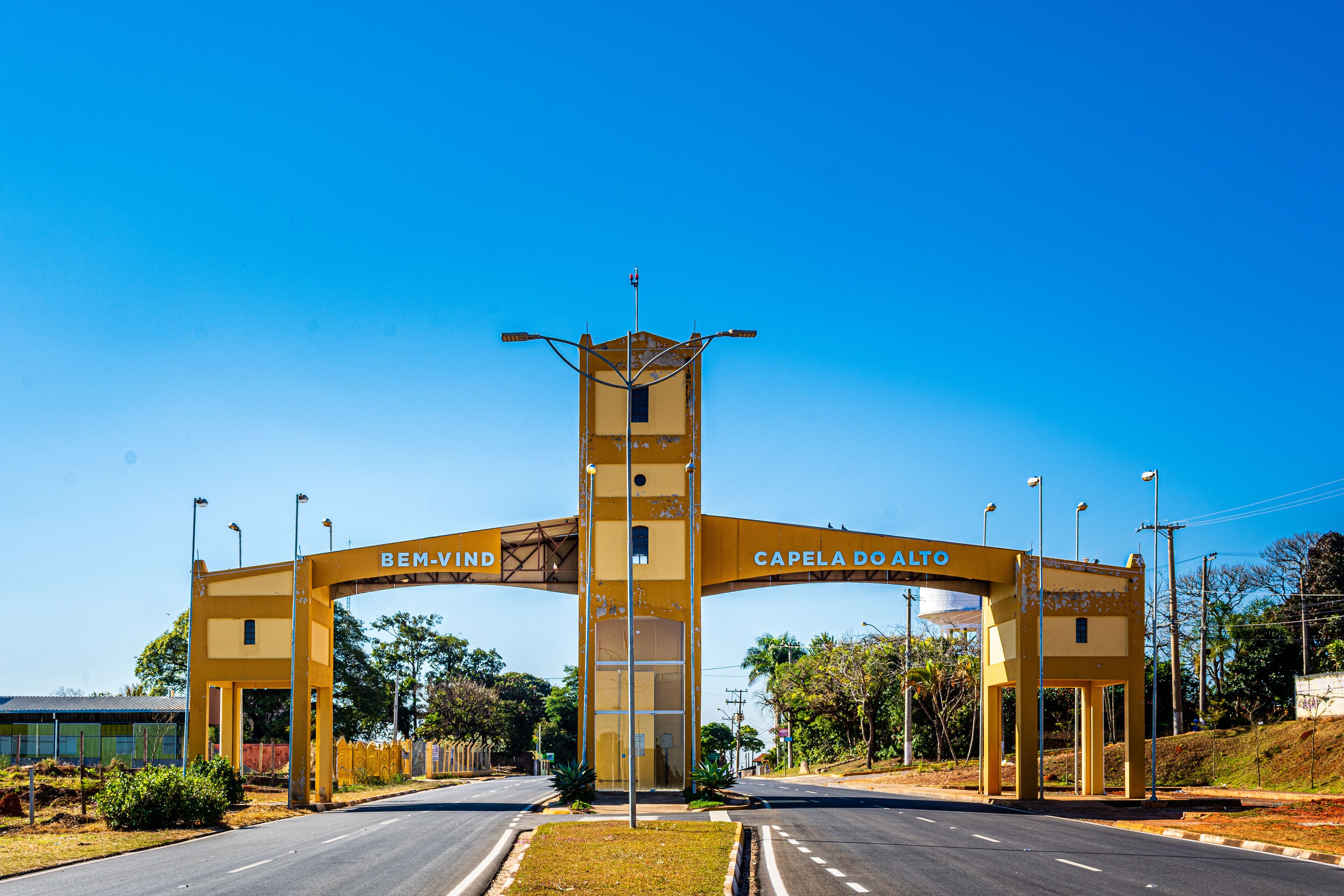 A yellow archway with "BEM-VIND" and "CAPELA DO ALTO" inscribed, leading to a road.