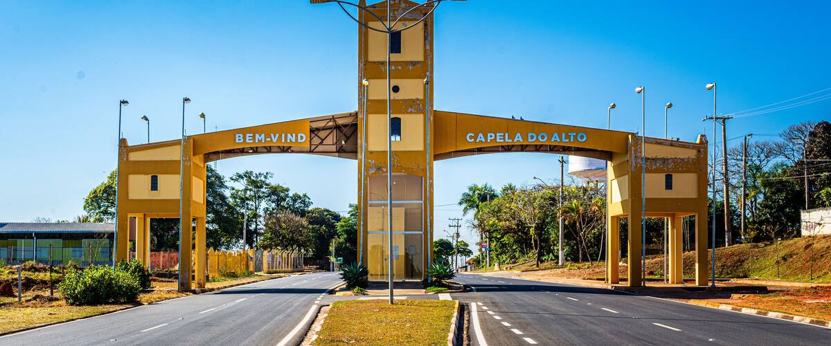 A yellow archway with "BEM-VIND" and "CAPELA DO ALTO" inscribed, leading to a road.