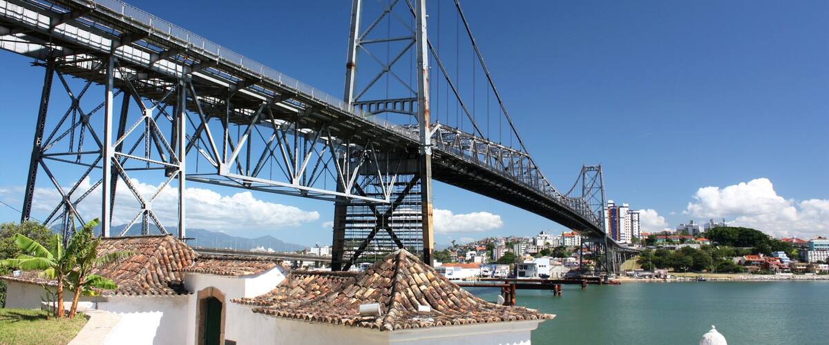 A large steel bridge spanning a river with a white building and palm trees nearby.