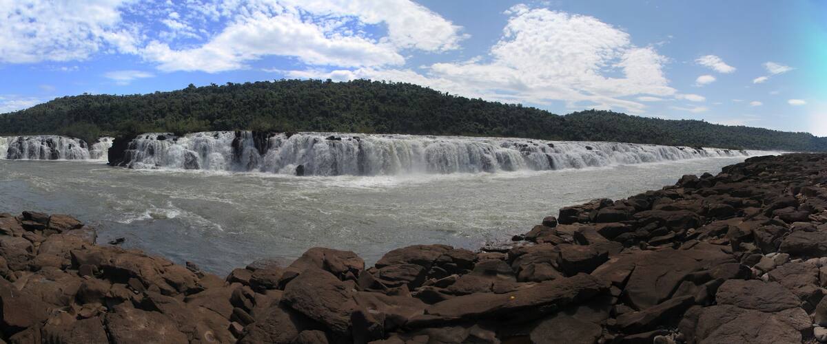 A waterfall cascading over a rocky riverbed, surrounded by a forested hillside.