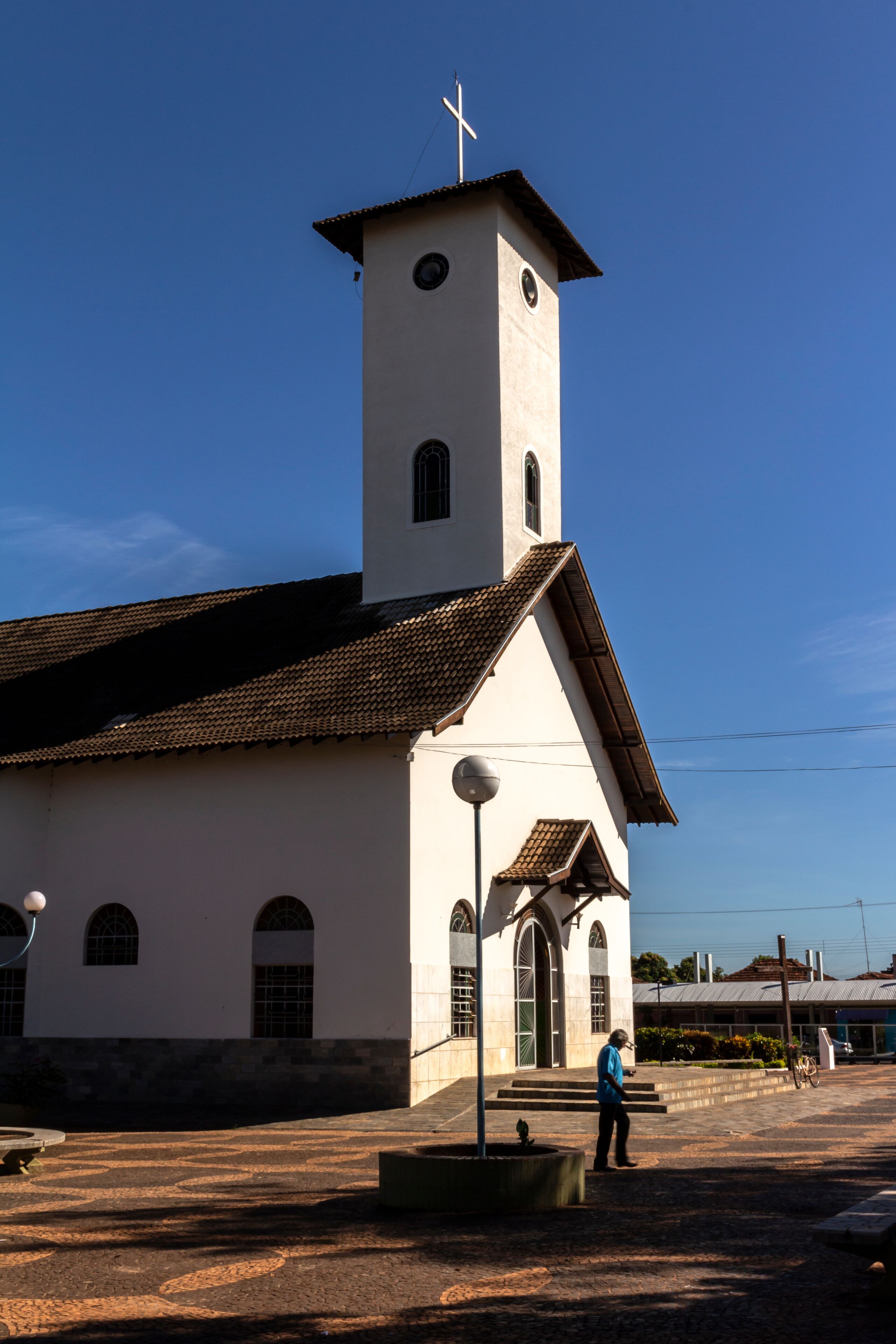 A white church with a bell tower and a cross on top, a person standing in front, and a clear blue sky.