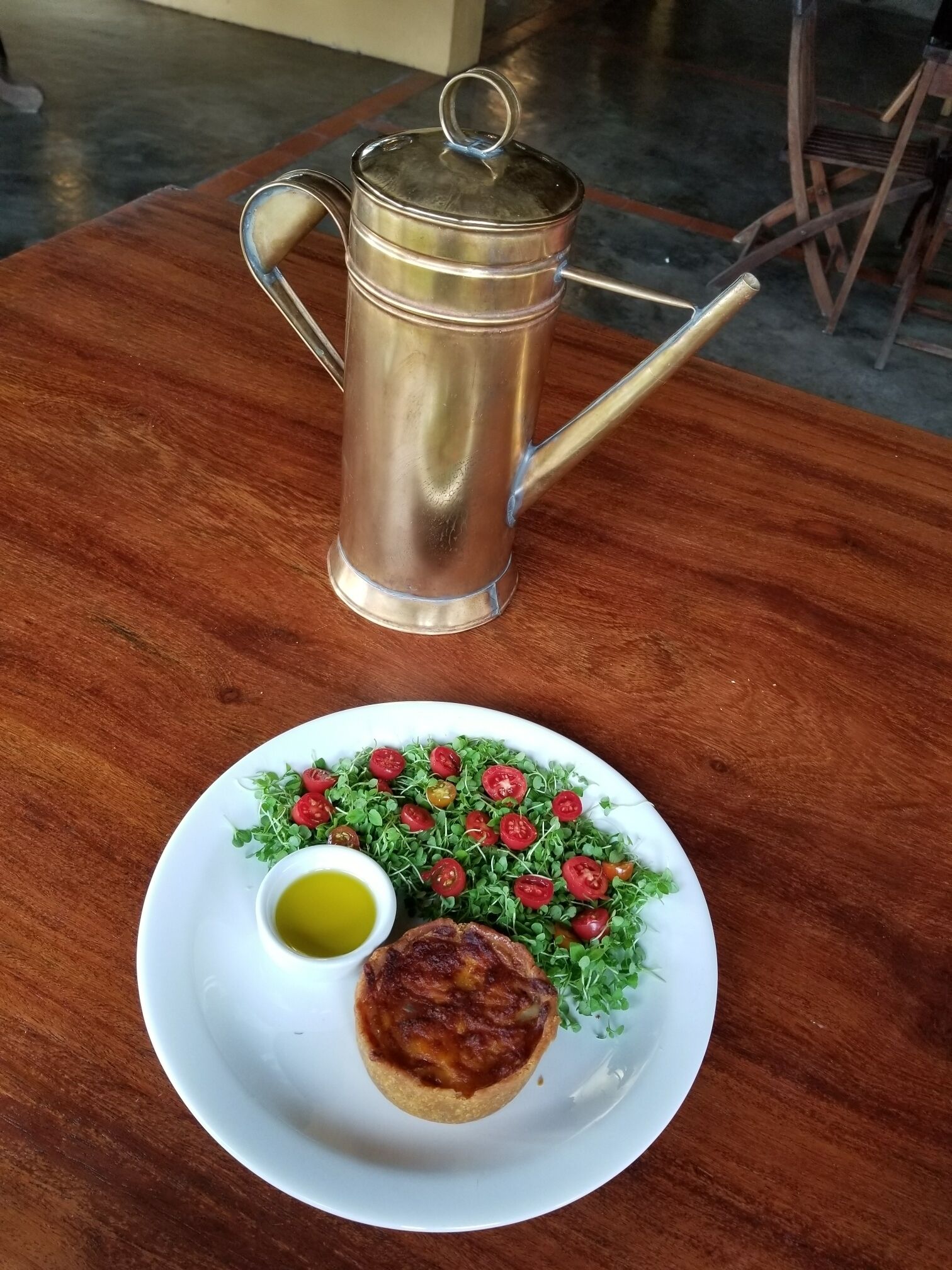 A metal coffee pot on a wooden table with a plate of food.