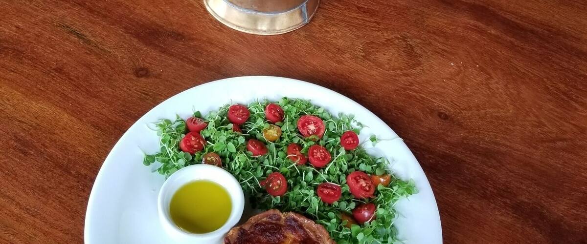 A metal coffee pot on a wooden table with a plate of food.