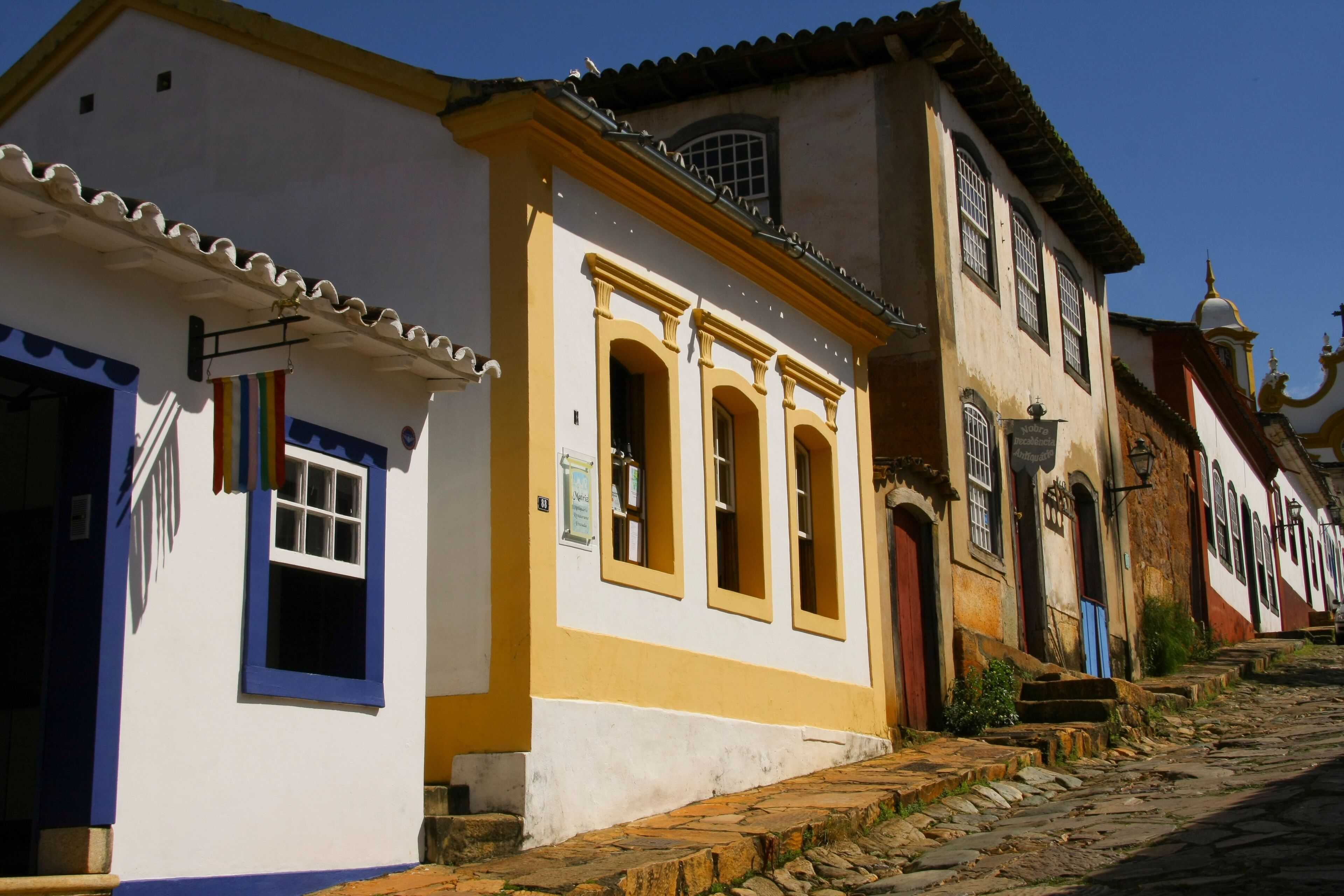 A cobblestone street lined with colorful buildings, including a white building with blue trim and a yellow building with arched windows.