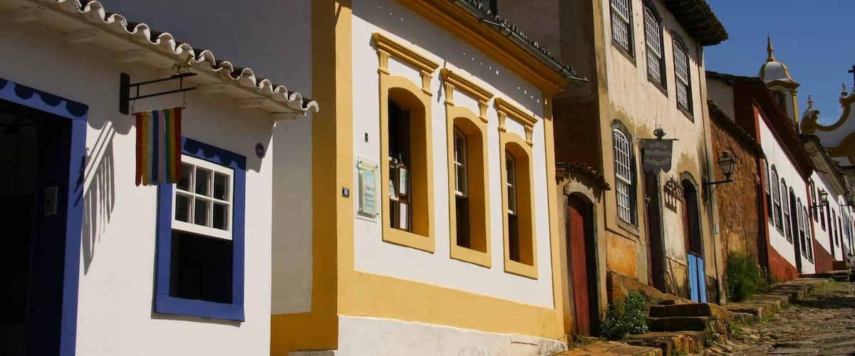 A cobblestone street lined with colorful buildings, including a white building with blue trim and a yellow building with arched windows.