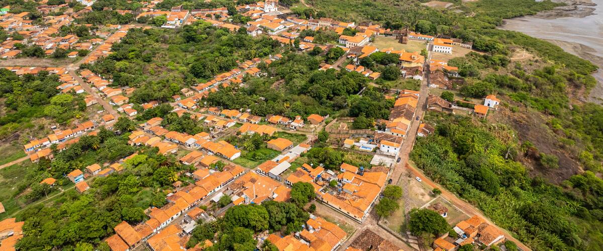 An aerial view of a coastal town with orange-roofed houses, green hills, and a beach.