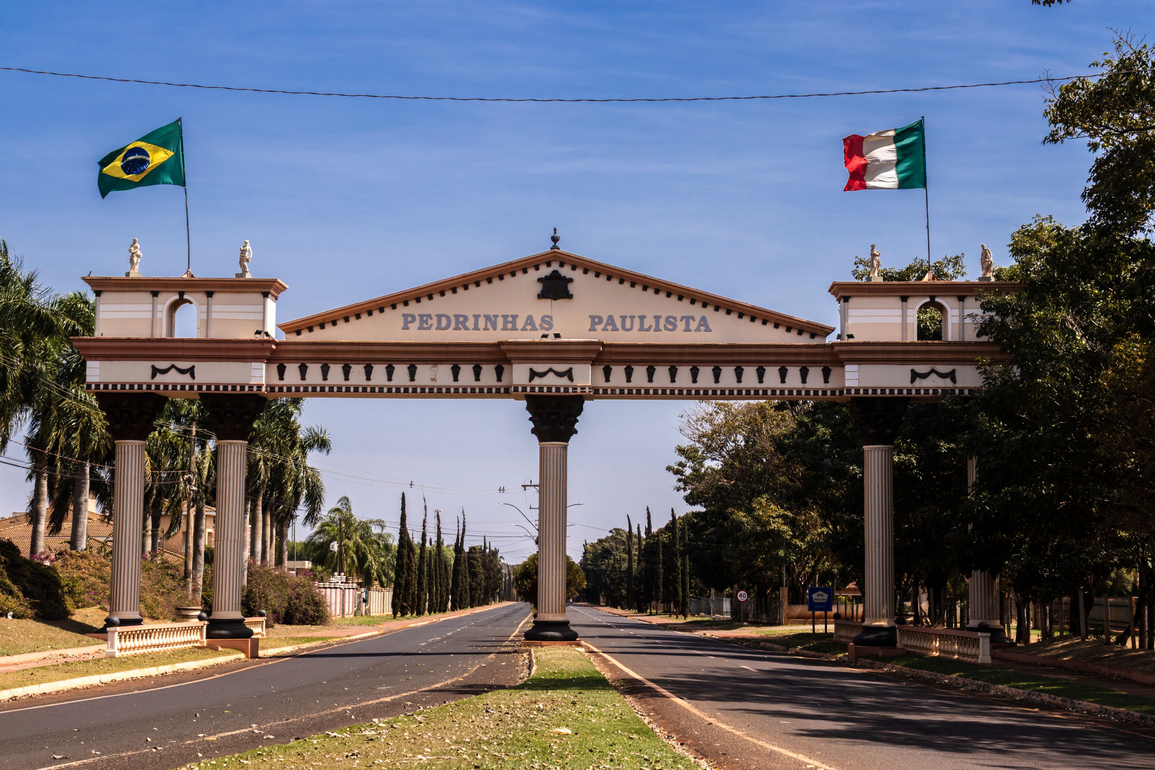 A road leading to a large archway with the inscription "PEDRINHAS PAULISTA" and flags of Brazil and Italy.