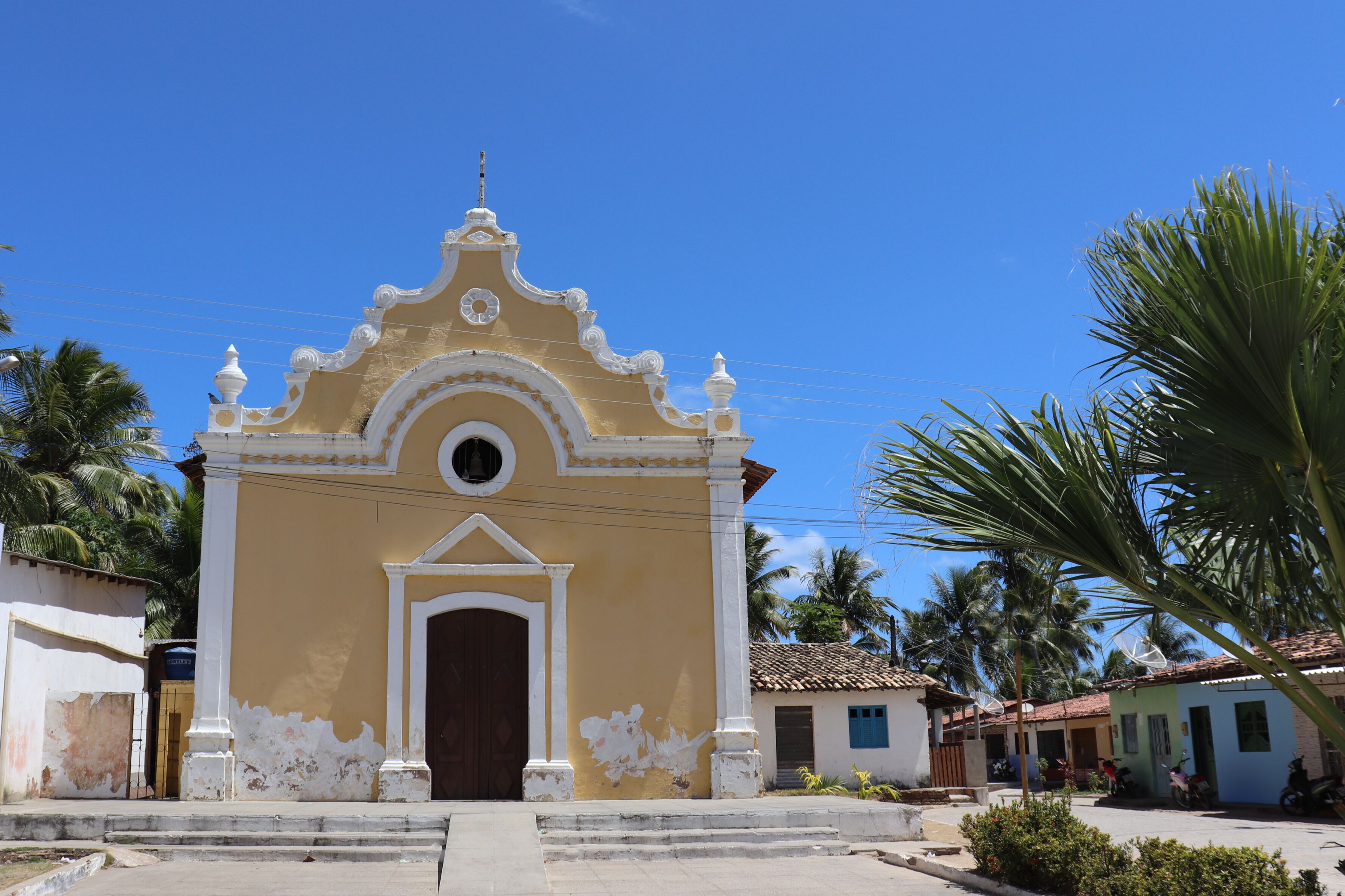 A yellow church with white trim and a brown door, surrounded by palm trees and small houses.