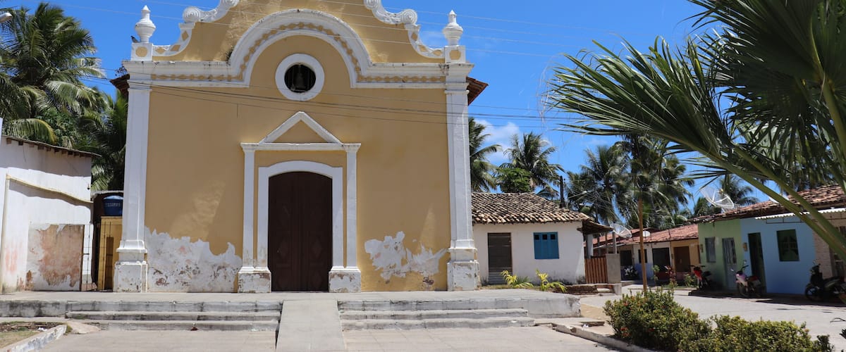 A yellow church with white trim and a brown door, surrounded by palm trees and small houses.