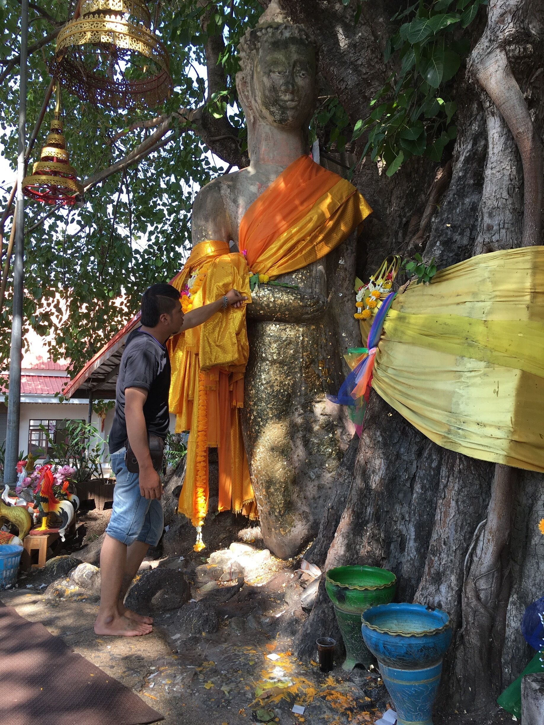 A large Buddha statue adorned with orange cloth and flowers, surrounded by trees and various decorative items.