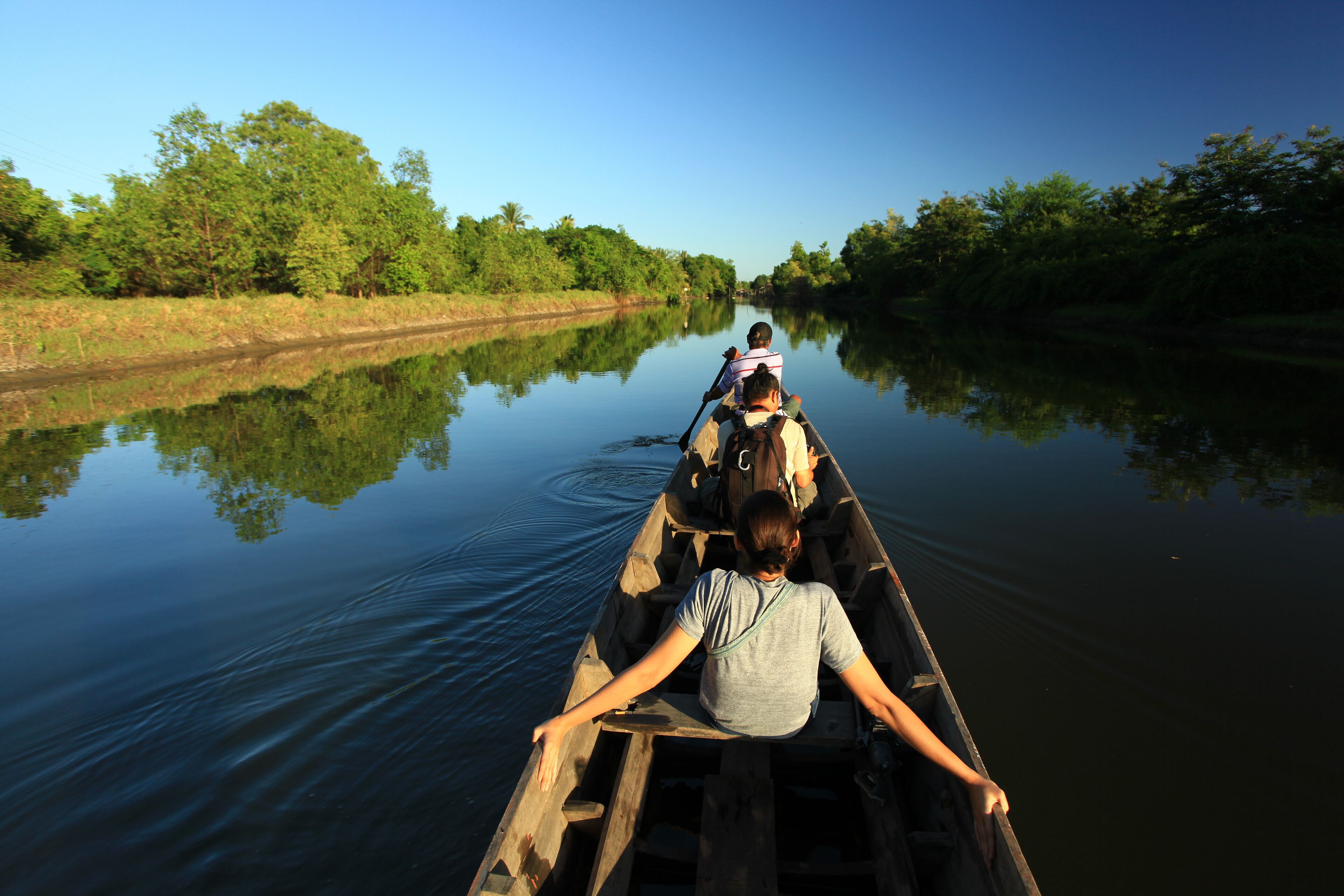 A narrow waterway flanked by dense greenery, with a wooden boat carrying two people.